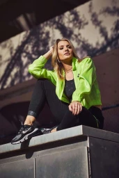 Woman wearing neon green jacket and black athletic pants seated on a concrete ledge, photographed by James Acomb.