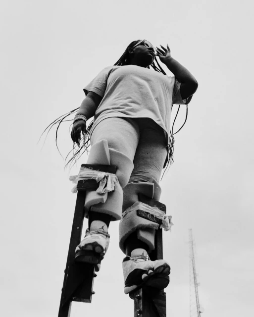 Low angle black and white shot of a woman with long braids wearing stilts, a t-shirt and pants against a bright overcast sky.