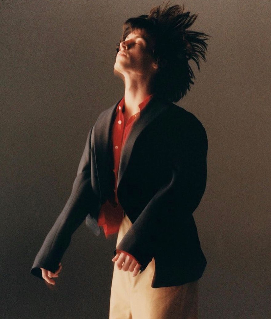Stylish individual with tousled hair, wearing a red shirt and blazer, in a fashion portrait against a neutral backdrop. 