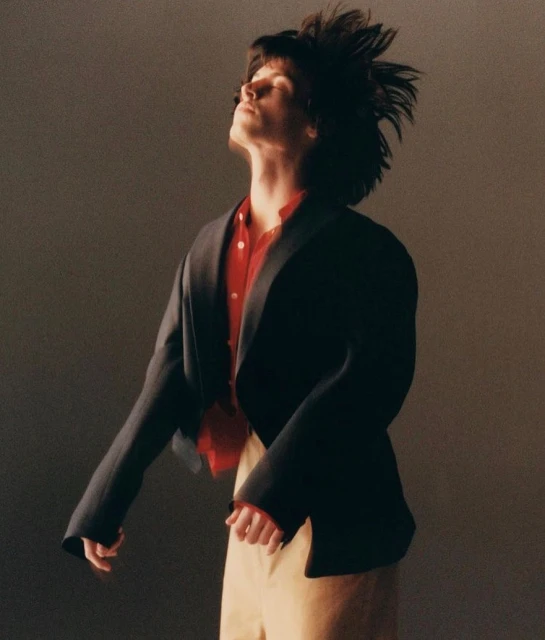 Stylish individual with tousled hair, wearing a red shirt and blazer, in a fashion portrait against a neutral backdrop. 