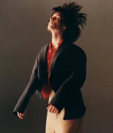 Stylish individual with tousled hair, wearing a red shirt and blazer, in a fashion portrait against a neutral backdrop. 