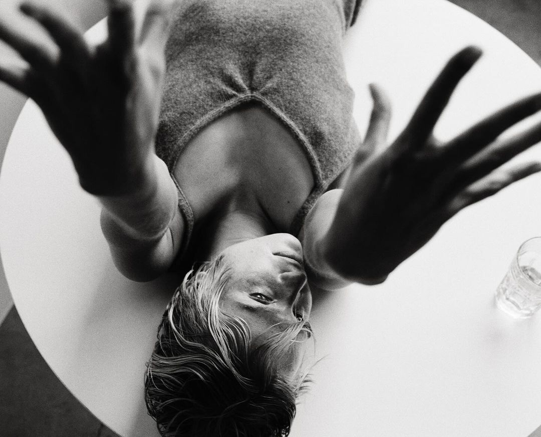 Overhead shot of a blonde woman lying on a white table, hands raised towards the camera, wearing a grey top, with a glass nearby.