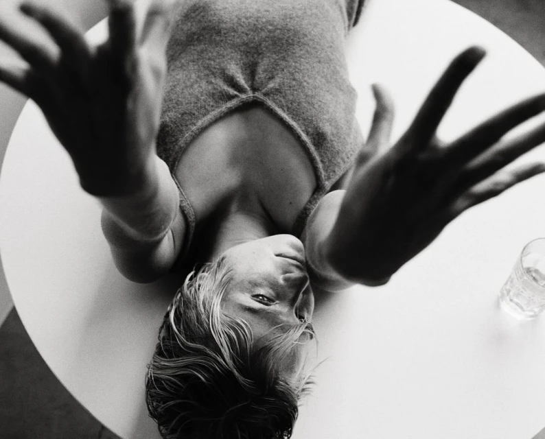 Overhead shot of a blonde woman lying on a white table, hands raised towards the camera, wearing a grey top, with a glass nearby.