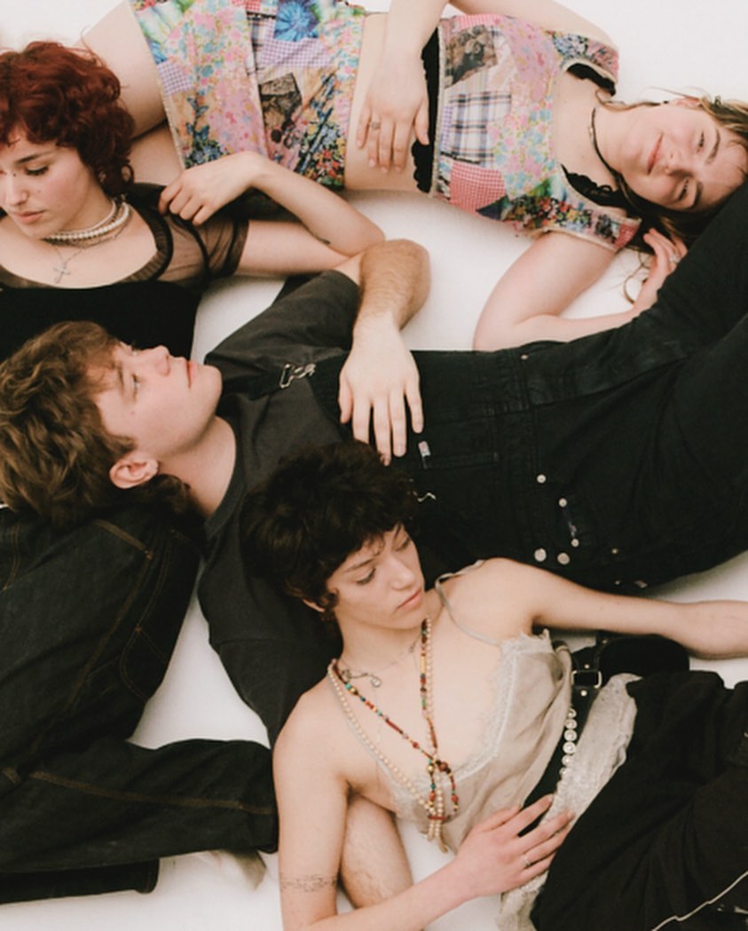 A group of five young adults laying together on a white surface, wearing a mix of dark and pastel-colored clothing in an indoor setting.