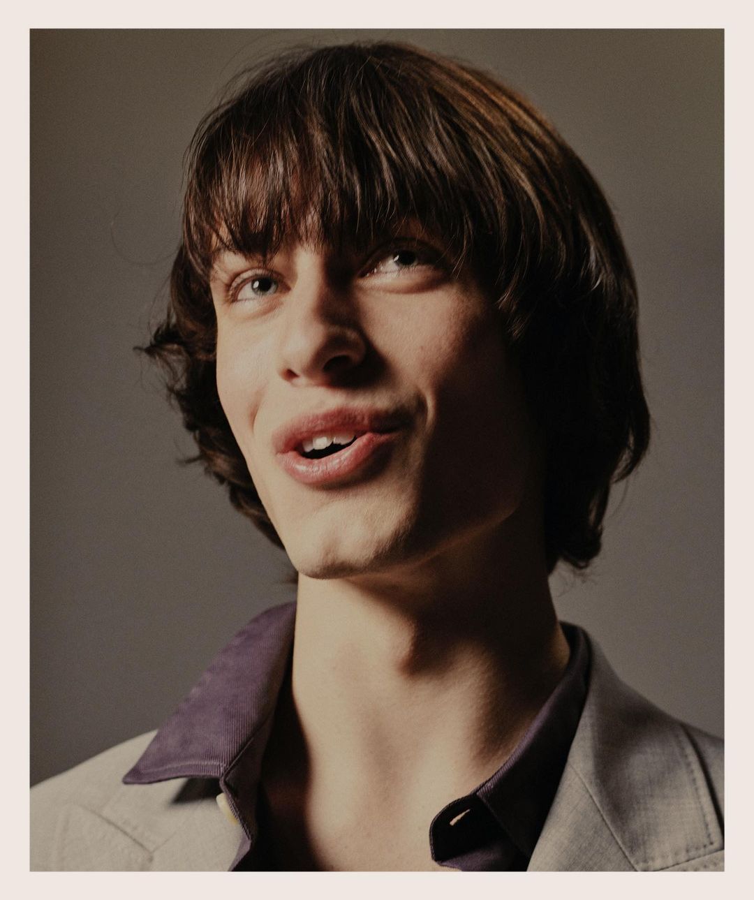 Close-up studio portrait of a young adult male with brown hair and light eyes, wearing a grey blazer over a purple collared shirt.