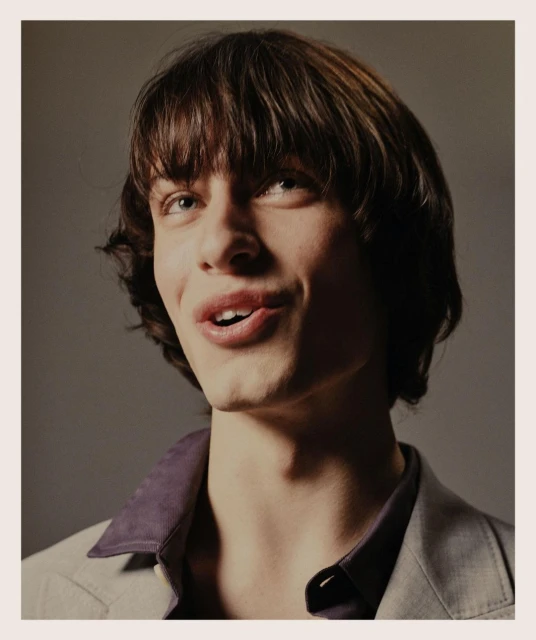 Close-up studio portrait of a young adult male with brown hair and light eyes, wearing a grey blazer over a purple collared shirt.