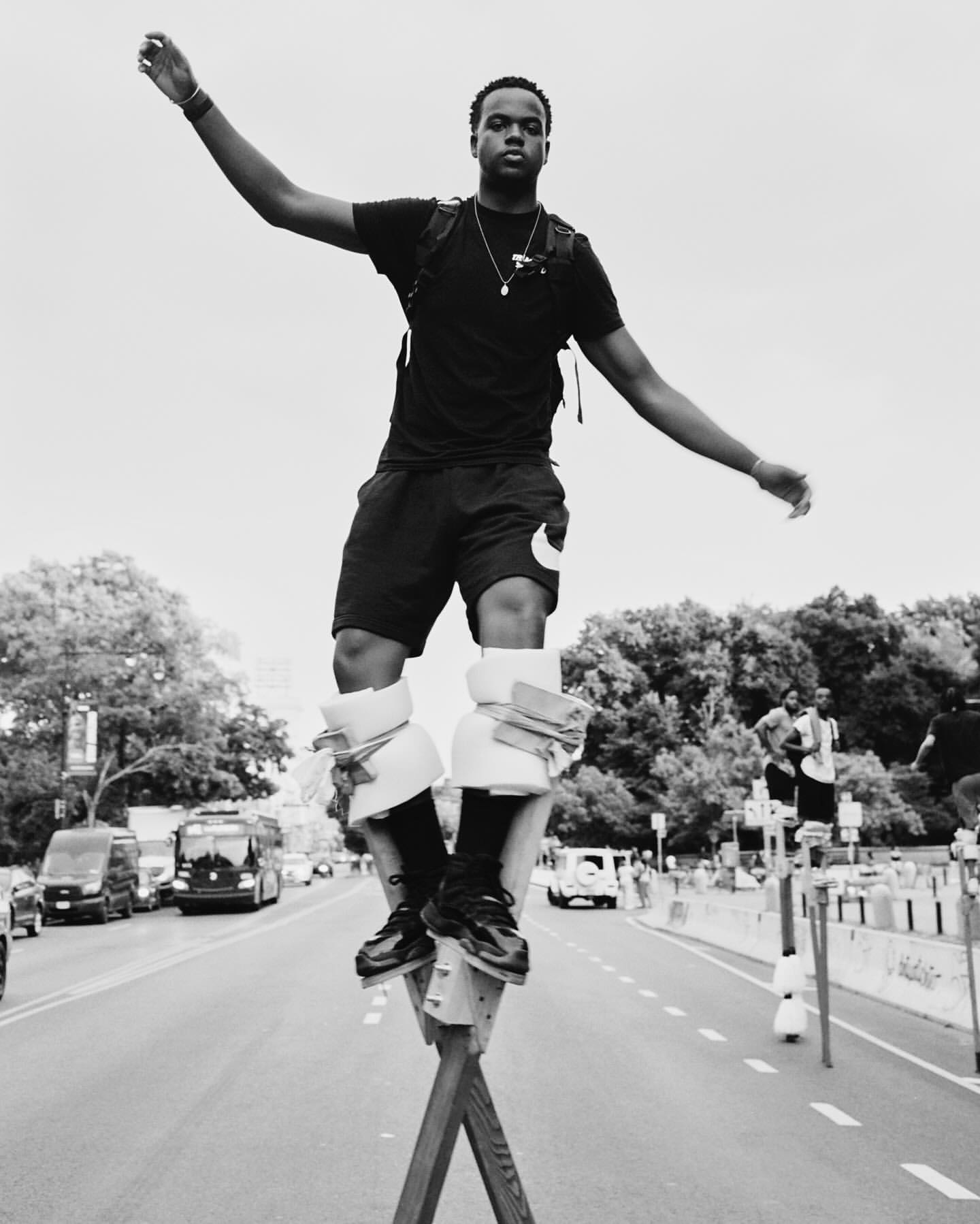 A confident young man balances on stilts in the street, wearing casual clothing, against a backdrop of trees and urban vehicles in black and white.