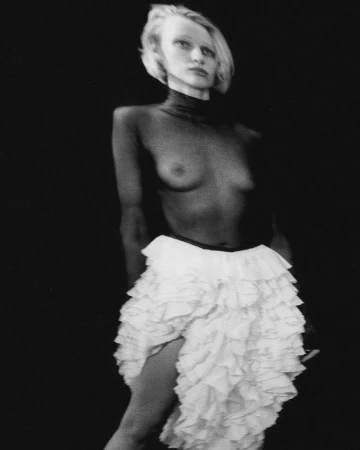 Striking black and white studio portrait of a topless adult female wearing a short, layered, frilly white skirt against a dark backdrop.