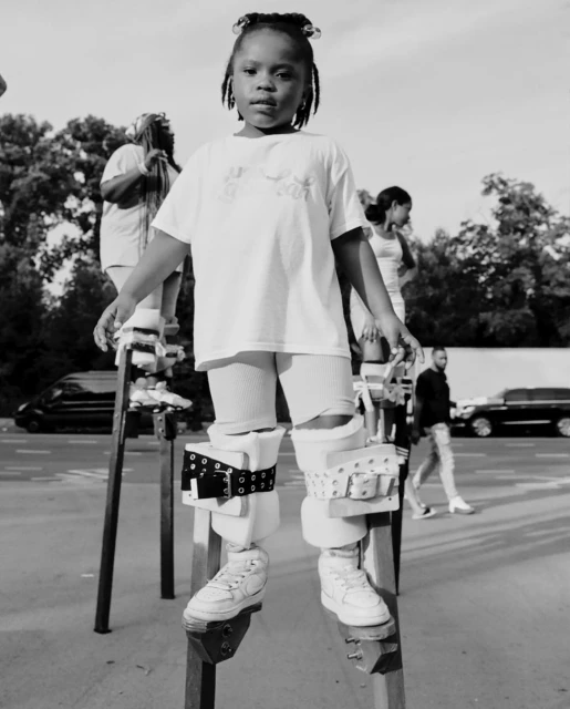 Black and white portrait of a young girl balancing on stilts outdoors on a street with other people and a black car in the background.