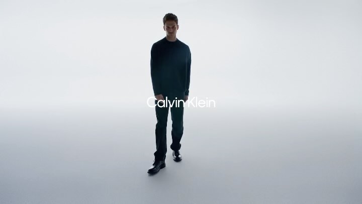 A man walks toward the camera wearing a black sweater, black pants, and black boots in a bright, high-key studio with copy overlay.