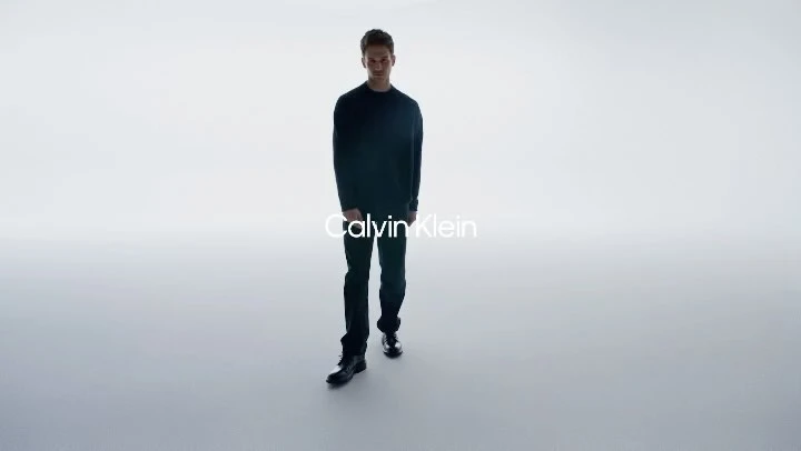 A man walks toward the camera wearing a black sweater, black pants, and black boots in a bright, high-key studio with copy overlay.