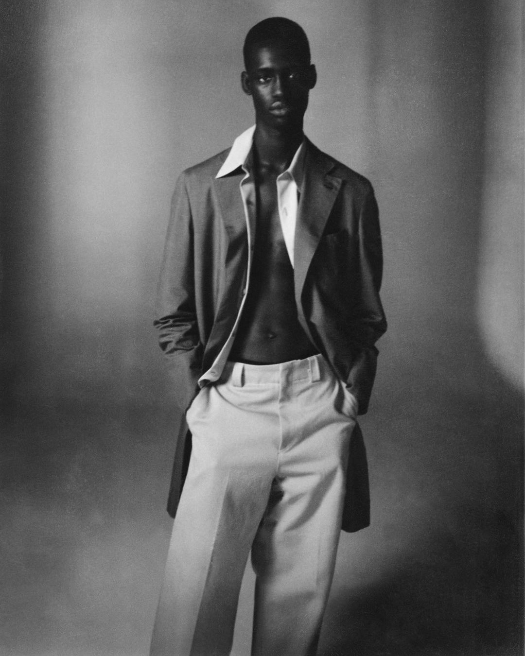 Monochrome portrait of a Black male model in a jacket and trousers, striking a pose against a neutral backdrop with hands in pockets.