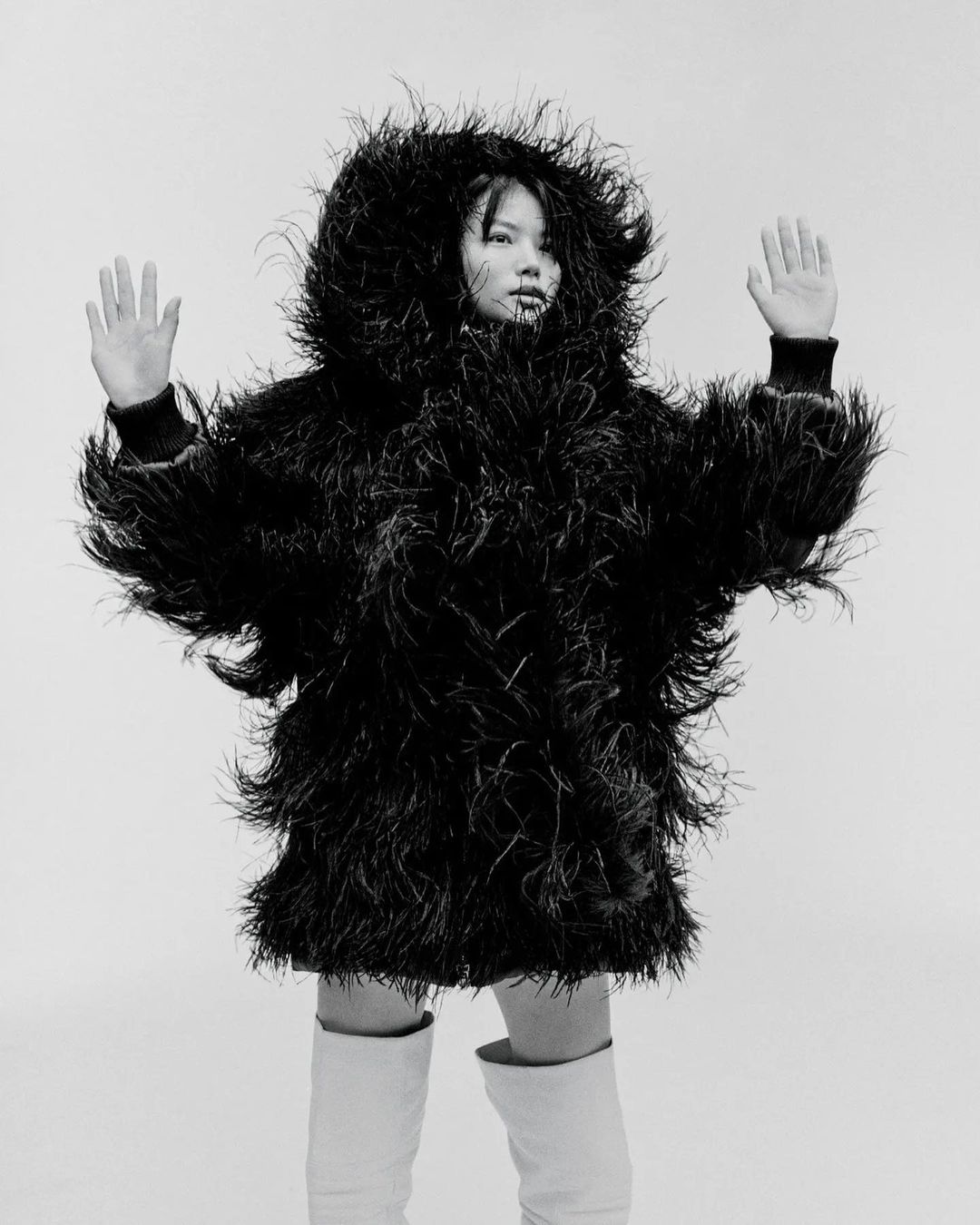 Monochrome fashion portrait of a model in a feathered jacket and white boots against a stark background, hands raised in a gesture.