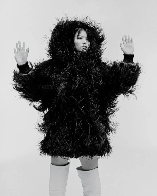 Monochrome fashion portrait of a model in a feathered jacket and white boots against a stark background, hands raised in a gesture.