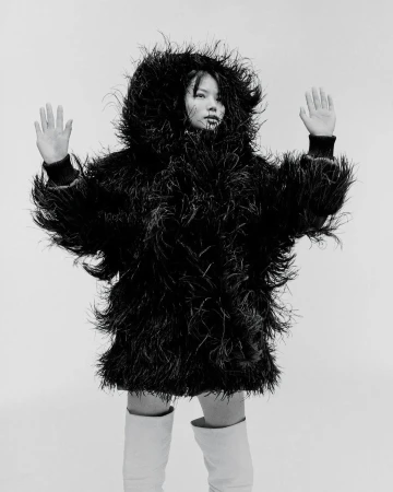 Monochrome fashion portrait of a model in a feathered jacket and white boots against a stark background, hands raised in a gesture.