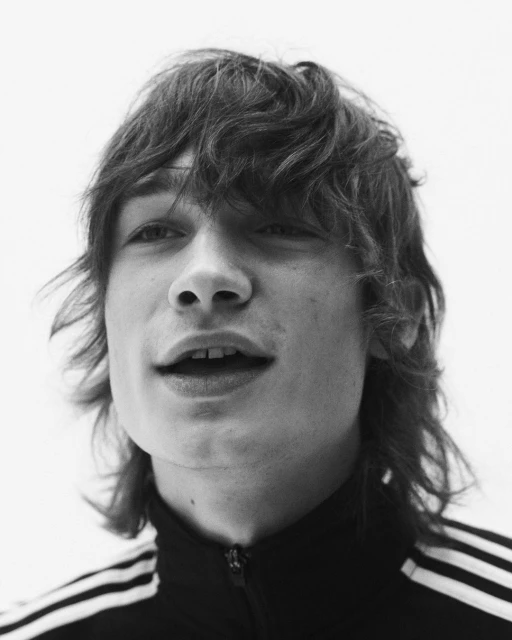 Black and white portrait of a young adult male with wavy hair wearing a zip up track jacket against a plain white backdrop.