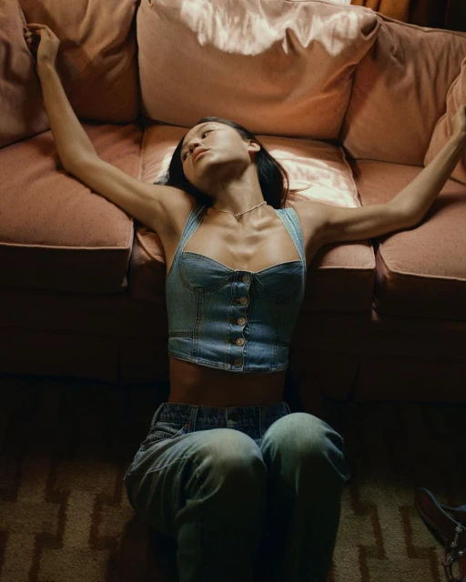 Young woman in a denim top and jeans lounges barefoot on the floor near a sofa. Indoor, natural lighting.