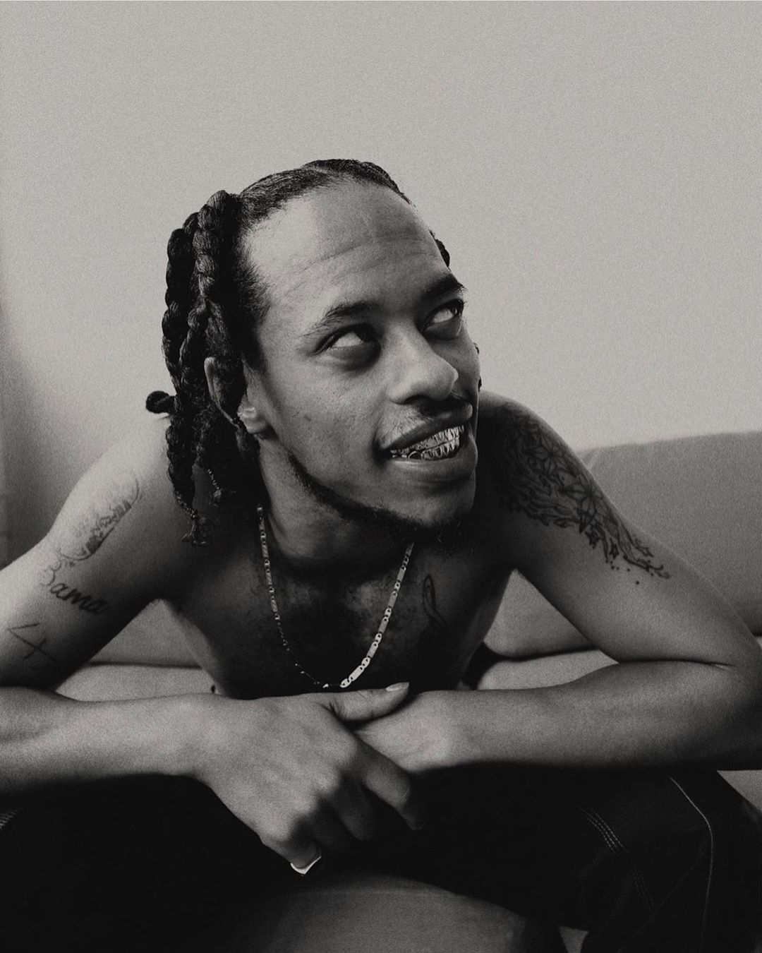 Monochrome portrait of Black man with braided hair, shirtless, gold teeth, and tattoos, seated indoors against a pale neutral background.