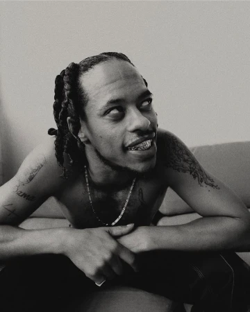 Monochrome portrait of Black man with braided hair, shirtless, gold teeth, and tattoos, seated indoors against a pale neutral background.