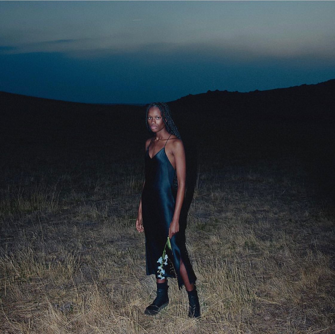 A young woman with braids poses in a black dress and boots in a field against a dark sky, holding flowers in a nighttime setting.