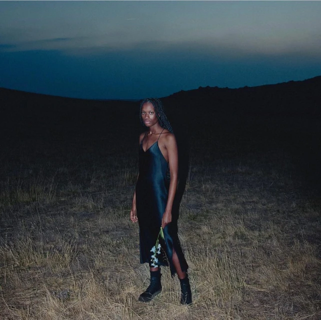 A young woman with braids poses in a black dress and boots in a field against a dark sky, holding flowers in a nighttime setting.