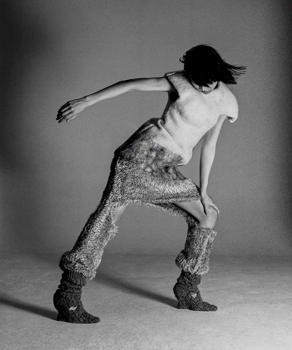 Monochrome fashion shot of a single slender female model in motion wearing a textured knit outfit in a photo studio with a grey backdrop.