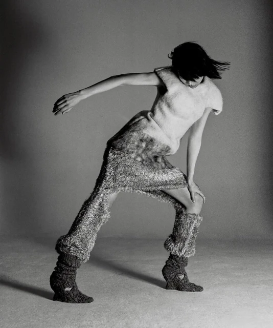Monochrome fashion shot of a single slender female model in motion wearing a textured knit outfit in a photo studio with a grey backdrop.