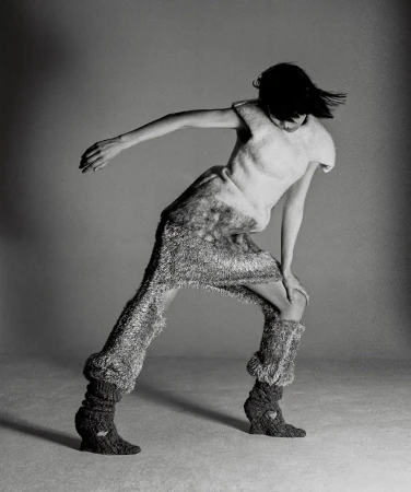 Monochrome fashion shot of a single slender female model in motion wearing a textured knit outfit in a photo studio with a grey backdrop.