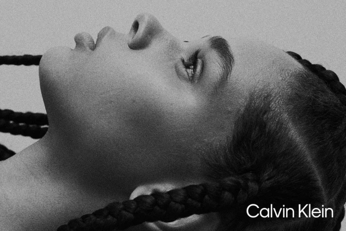 Close-up black and white portrait of a person with braided hair looking up against a light gray background, featuring the Calvin Klein logo.