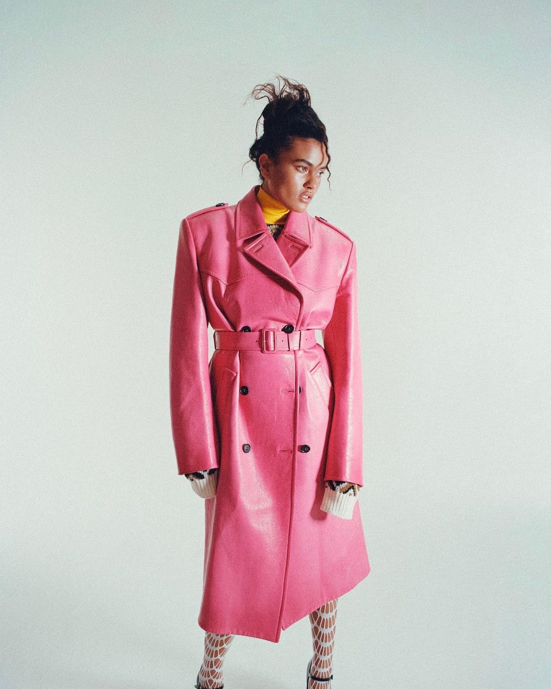 Woman in a vibrant pink trench coat, paired with patterned stockings and heels, striking a pose against a plain white background in studio setting.