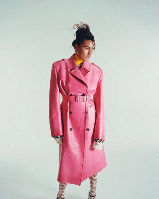 Woman in a vibrant pink trench coat, paired with patterned stockings and heels, striking a pose against a plain white background in studio setting.