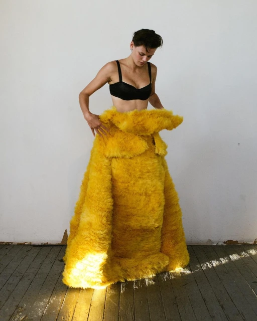 Woman in a black bra standing indoors wearing a large yellow faux fur skirt, posing in front of a white wall with a wooden floor.