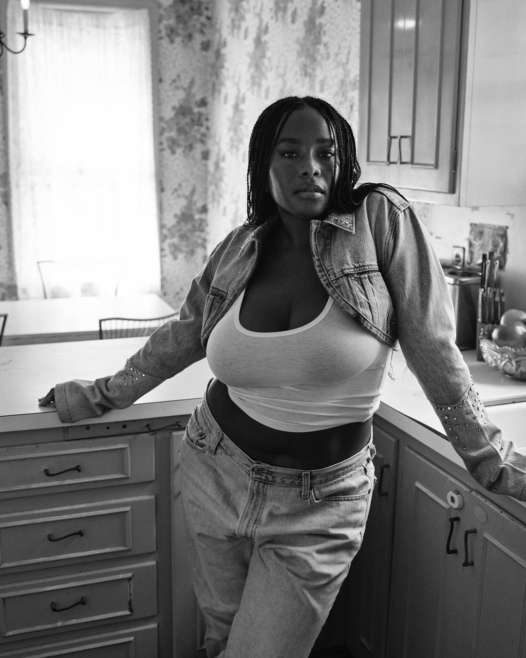 Monochrome portrait of a woman with braided hair in jeans and a denim jacket posing in a kitchen setting with floral wallpaper.
