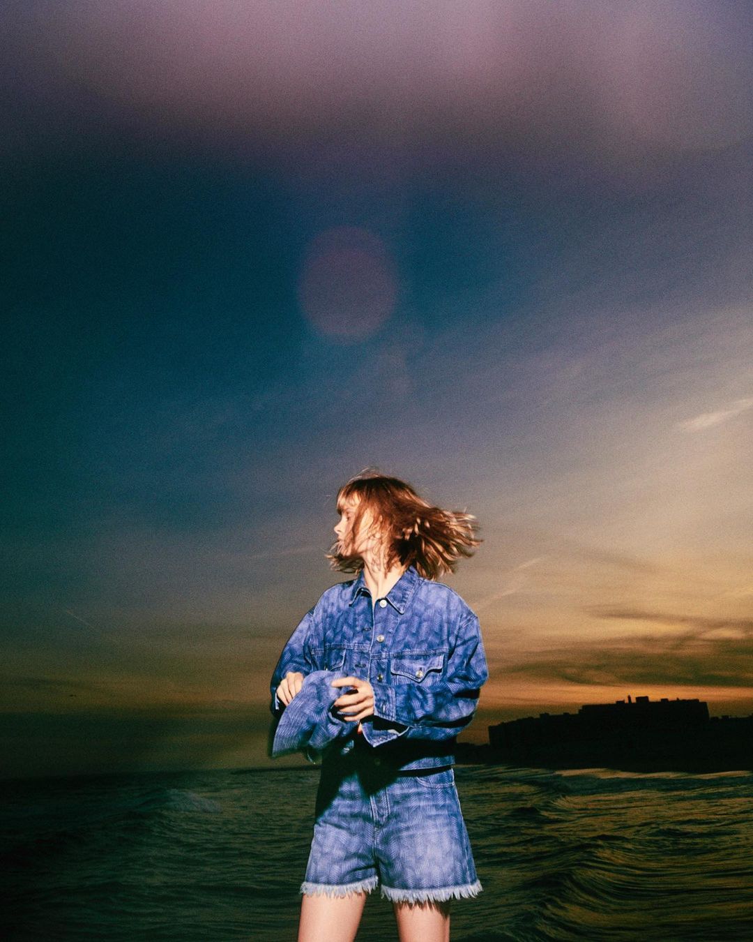 A stylish young woman in a denim jacket and shorts stands on a beach with a colorful sunset sky and the ocean in the background.