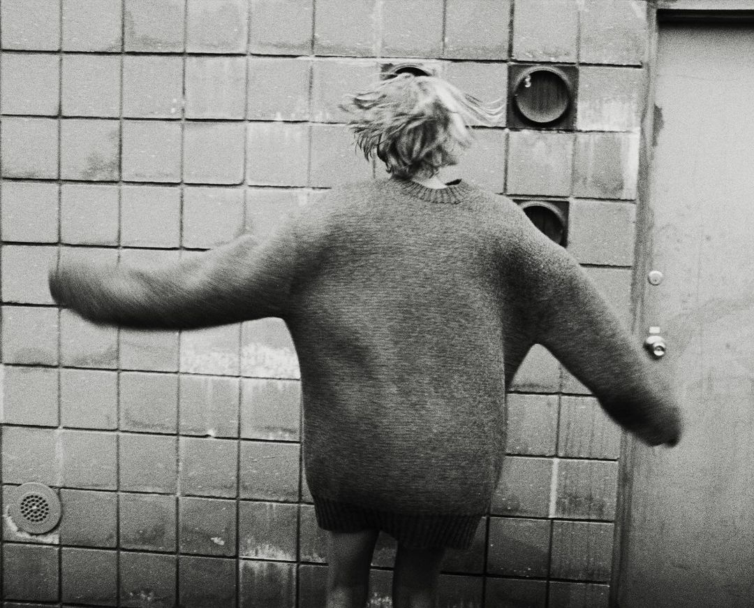 Person in oversized sweater stands in front of tiled wall with vents. Back view of individual with short blonde hair, black and white.