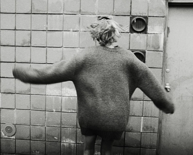 Person in oversized sweater stands in front of tiled wall with vents. Back view of individual with short blonde hair, black and white.