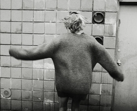 Person in oversized sweater stands in front of tiled wall with vents. Back view of individual with short blonde hair, black and white.