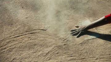 A hand reaches out to touch sand on a sunlit beach, creating ripples and patterns in the sand with a sense of texture and movement.