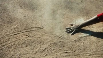 A hand reaches out to touch sand on a sunlit beach, creating ripples and patterns in the sand with a sense of texture and movement.