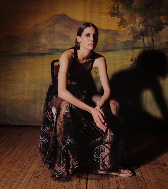 Fashionable woman poses seated in a dark lace gown with an painted mountain landscape backdrop indoors, showcasing elegance and style.