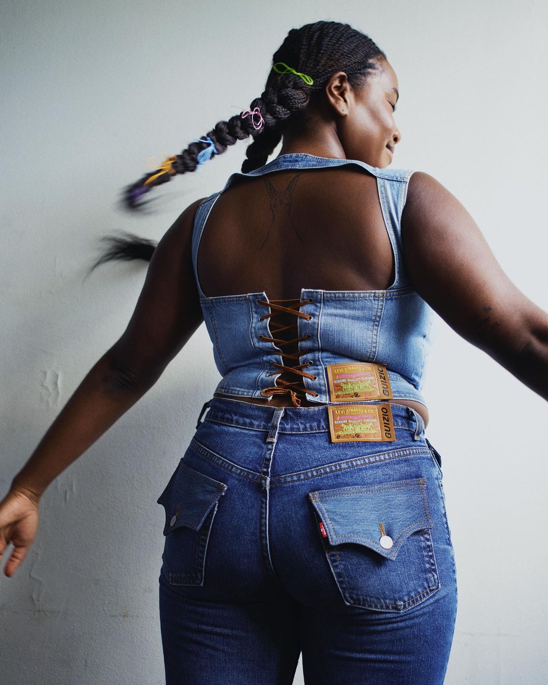 A Black woman in a denim corset top and jeans poses with outstretched arms against a plain background in a fashion shot.