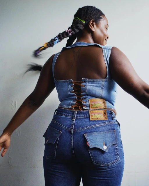 A Black woman in a denim corset top and jeans poses with outstretched arms against a plain background in a fashion shot.