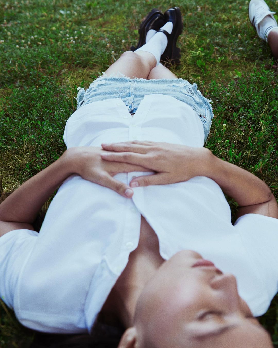 Adult female relaxes on green grass wearing a white top and jean skirt with black shoes and white socks, shot from an eye-level angle.