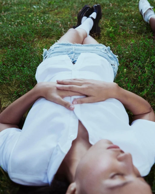 Adult female relaxes on green grass wearing a white top and jean skirt with black shoes and white socks, shot from an eye-level angle.