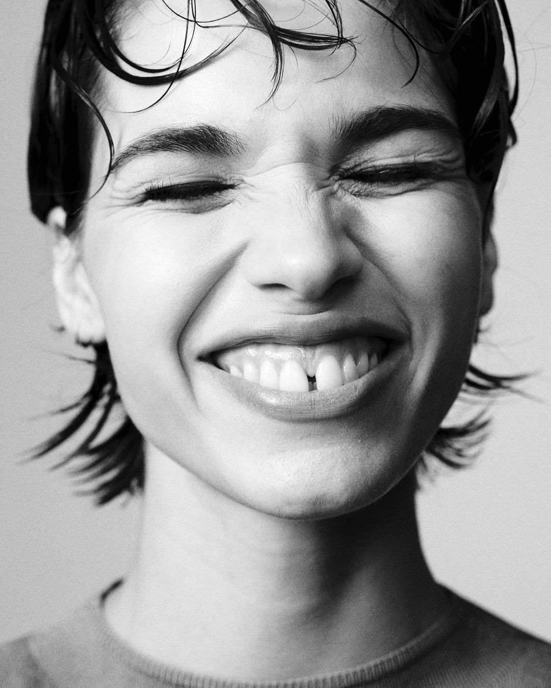 Black and white portrait of a young woman smiling with her eyes closed. She has short, dark, wet hair and is wearing a crew neck sweater.