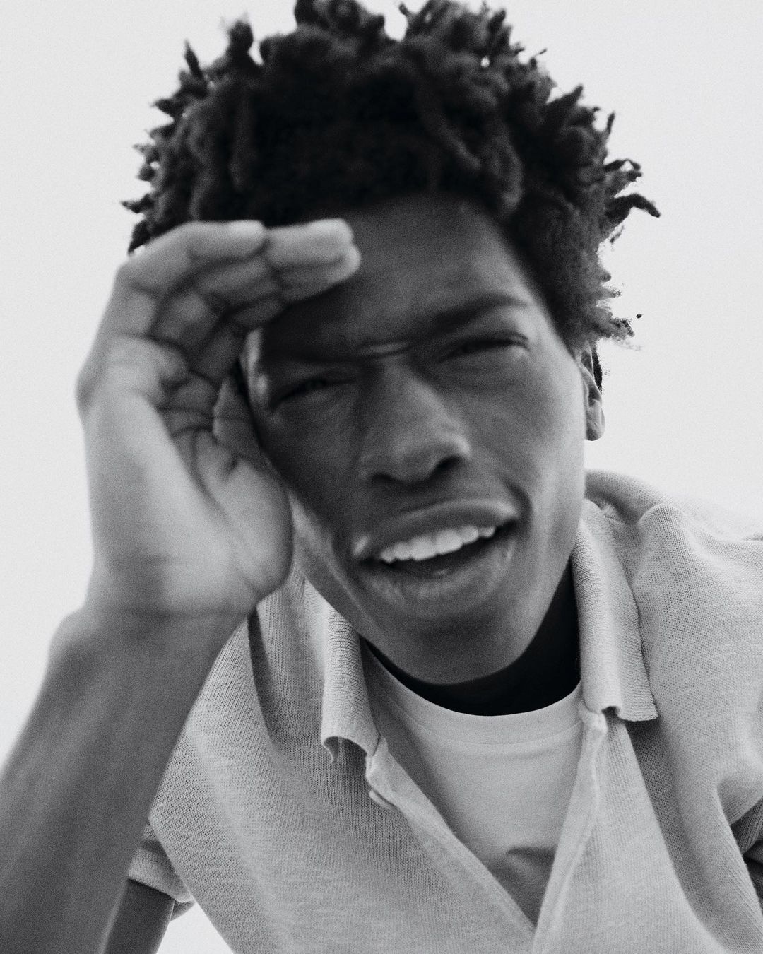 Monochrome close-up of a Black adult male with dreadlocks wearing a polo shirt, shielding eyes with hand against bright background.