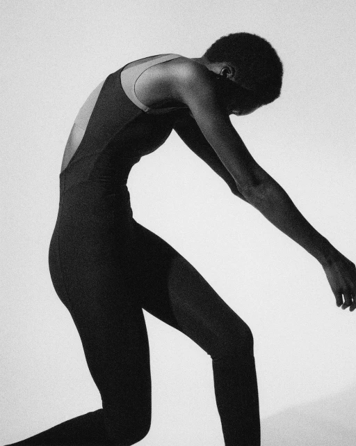 Slim female model in a dark bodysuit with an open back bends over in a minimalist studio setting, captured in a monochrome aesthetic.