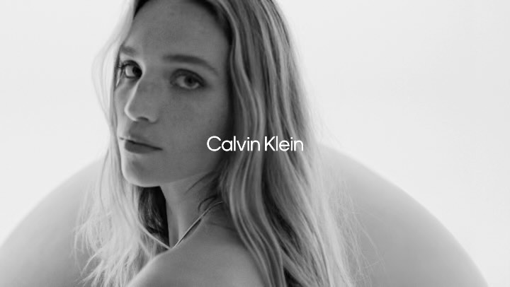 Monochrome portrait of a blonde-haired woman gazing over her shoulder against a bright white backdrop, featuring the Calvin Klein logo.