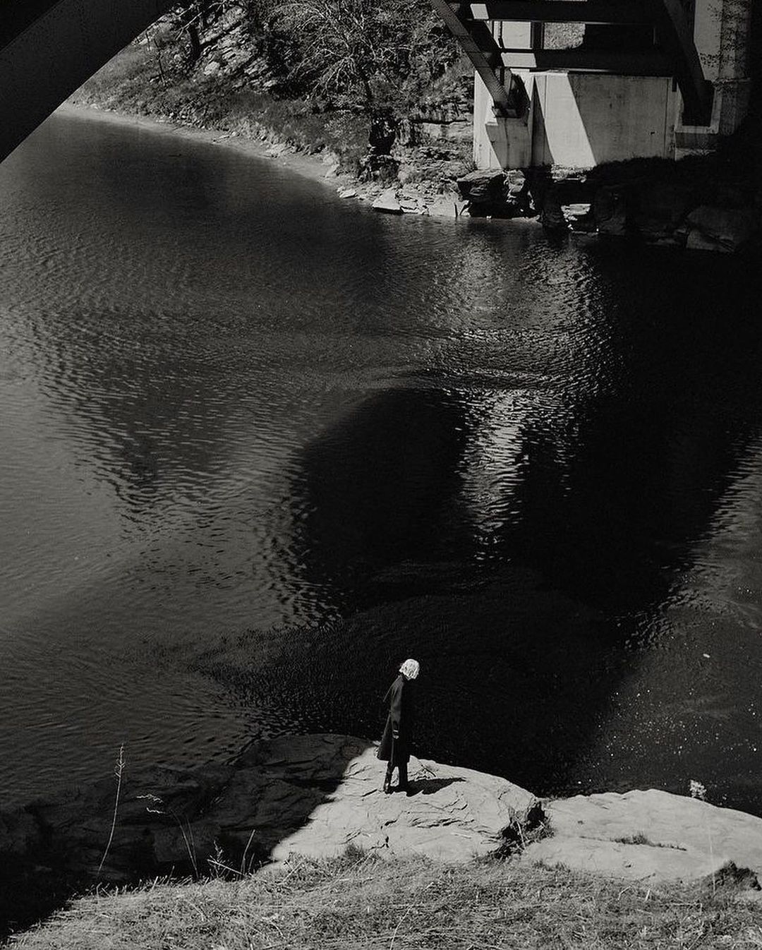 Monochromatic photograph of a person standing on a rock by a river under a bridge, creating a reflective shadow in the water.