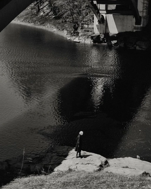 Monochromatic photograph of a person standing on a rock by a river under a bridge, creating a reflective shadow in the water.